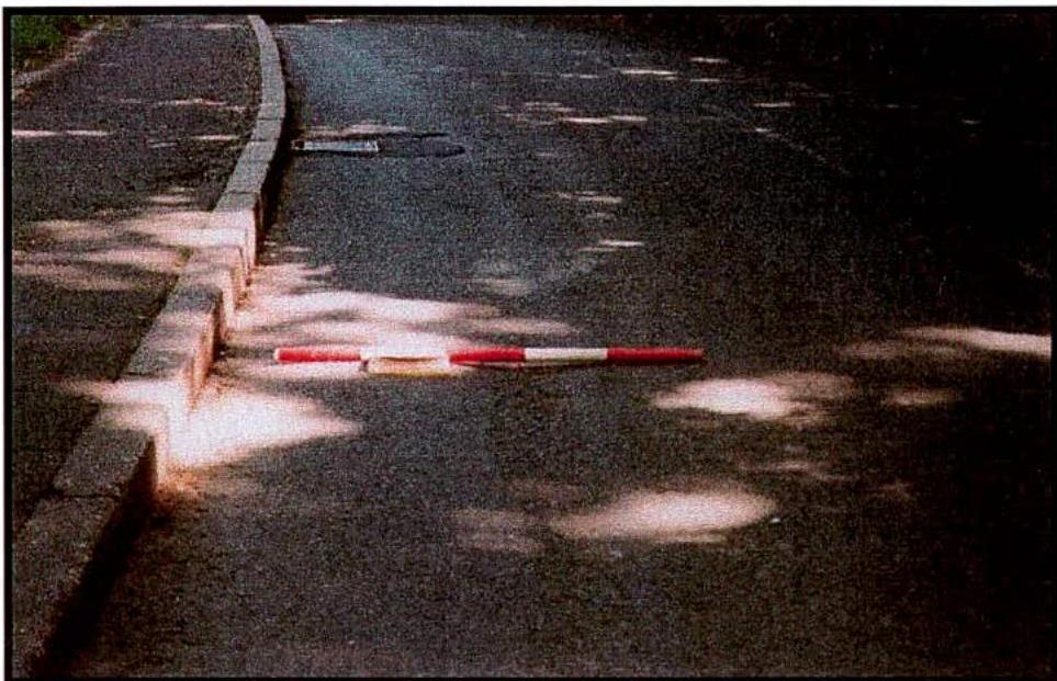 A grainy photograph showing a section of asphalt road with a concrete curb on the left and a red-and-white striped marker pole lying on the ground.
