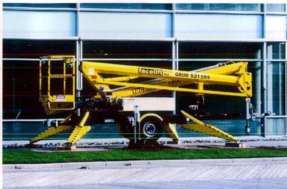 A photograph showing a yellow cherry picker parked on a paved area in front of a modern building with large glass windows.