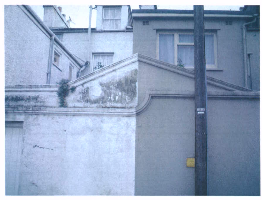 A photograph showing the exterior of a property featuring a garage door on the left and a pedimented wall section on the right, with a utility pole in the foreground.