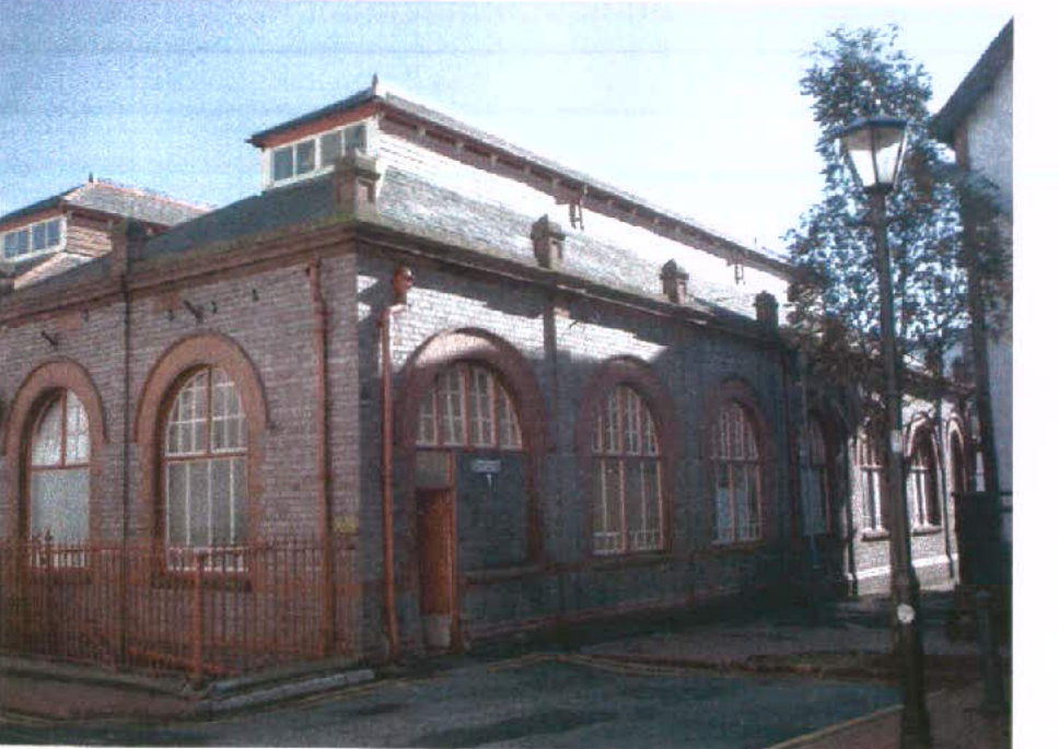 A photograph showing the exterior of a two-story brick building with large arched windows and a slate roof, likely an existing condition shot for a lighting application.