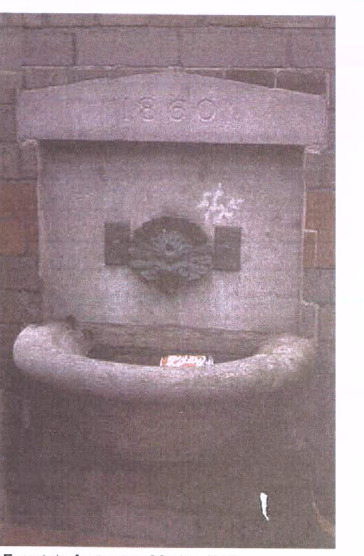 A photograph of a historic stone drinking fountain or trough built into a brick wall, featuring the date 1860 carved into the stone lintel.