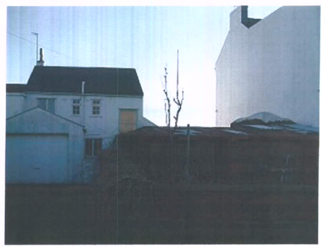A grainy photograph showing an existing white building with a dark roof on the left and a large white wall on the right, separated by a dark slope or vegetation.
