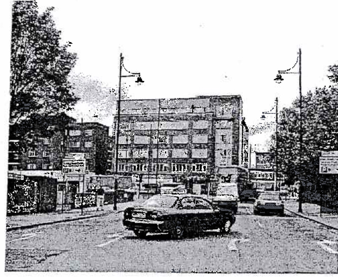 A grainy black and white photograph showing a street scene with cars, street lamps, and multi-story buildings in the background.