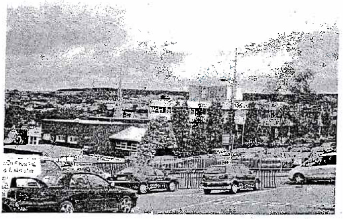A grainy black and white photograph showing a street scene with parked cars, a fence, and buildings in the background.