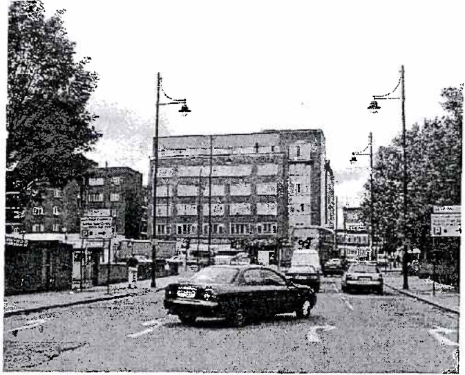 A grainy black and white photograph depicting a street scene with cars, streetlights, and multi-story buildings in the background.