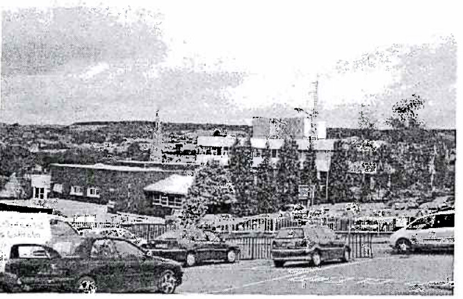 A grainy black and white photograph showing a parking area with cars in the foreground and a landscape with buildings and trees in the background. A tall, thin monopole structure is visible in the distance, likely the...