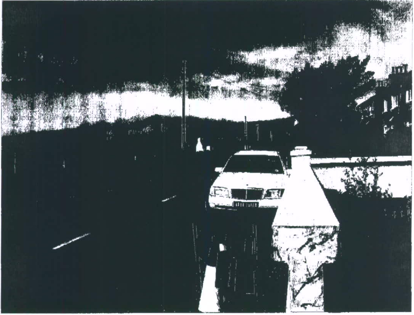 A high-contrast, grainy black and white photograph showing a car parked on a driveway next to a stone wall and road.