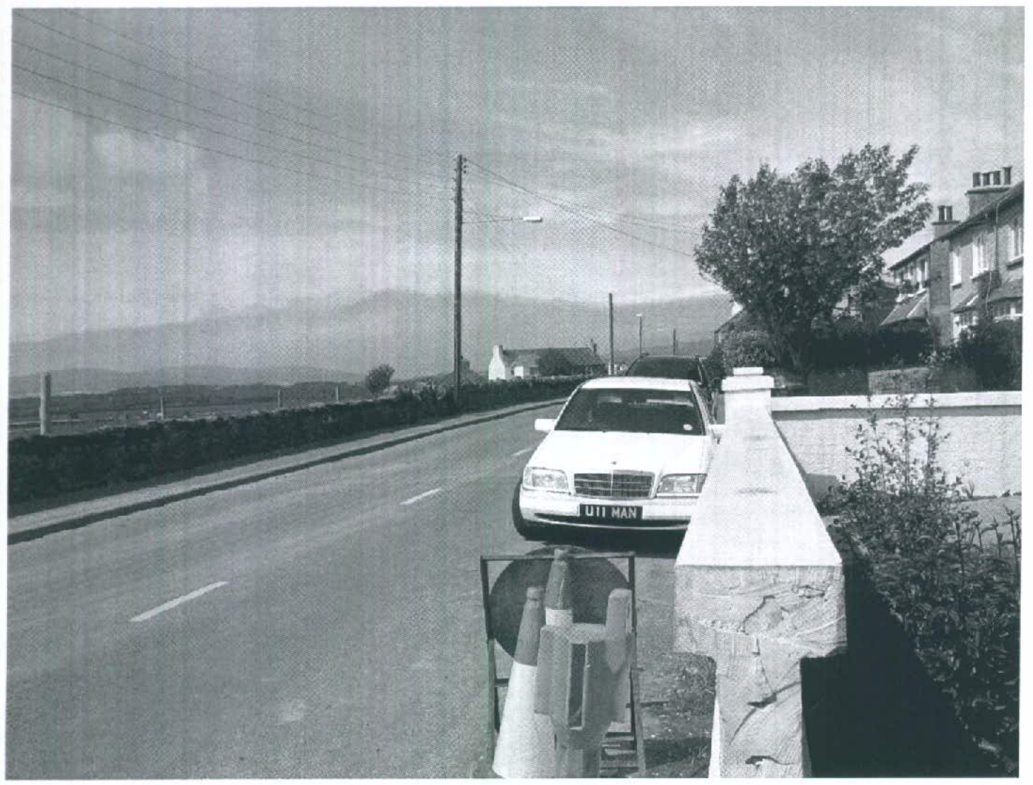 A black and white photograph showing a street scene with a white car parked near a stone wall entrance, with traffic cones in the foreground and hills in the distance.