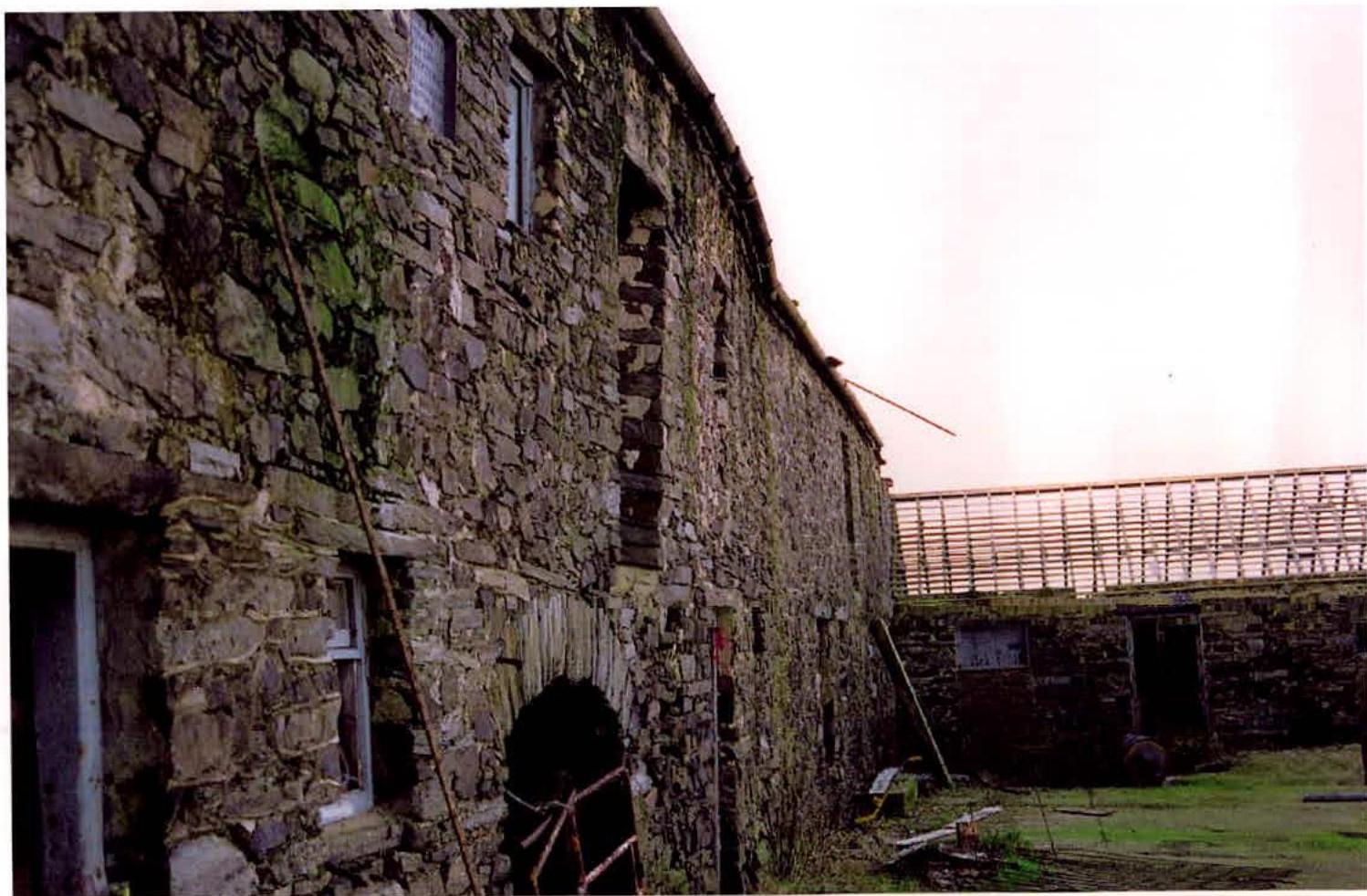 A photograph showing the exterior of a large, old stone barn with rough-hewn walls and window openings.