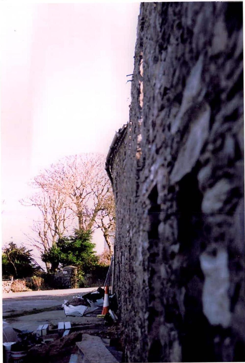 A close-up photograph showing the rough stone wall of a building, likely a barn, with construction debris and a traffic cone at the base.