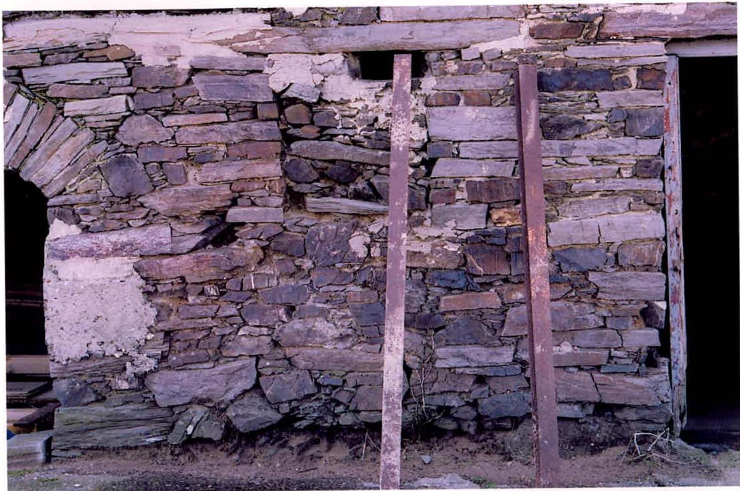 A close-up photograph of a weathered stone wall, likely part of a barn, supported by two vertical metal beams. The image captures the texture of the stonework and openings, indicating structural work or renovation.