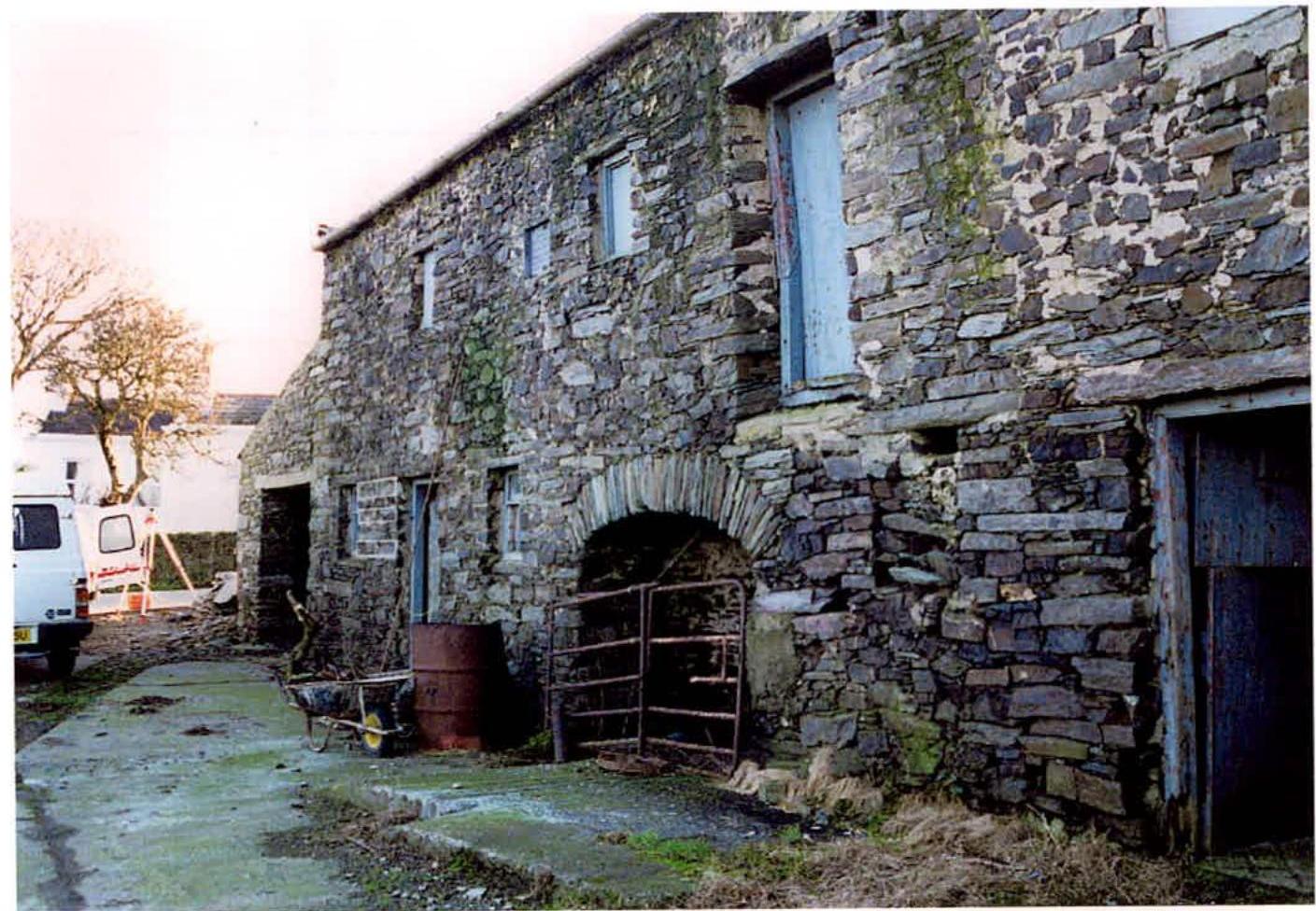 A photograph showing the exterior of a weathered stone barn with boarded windows and a large arched cart entrance, situated in a rural setting with a white van parked nearby.