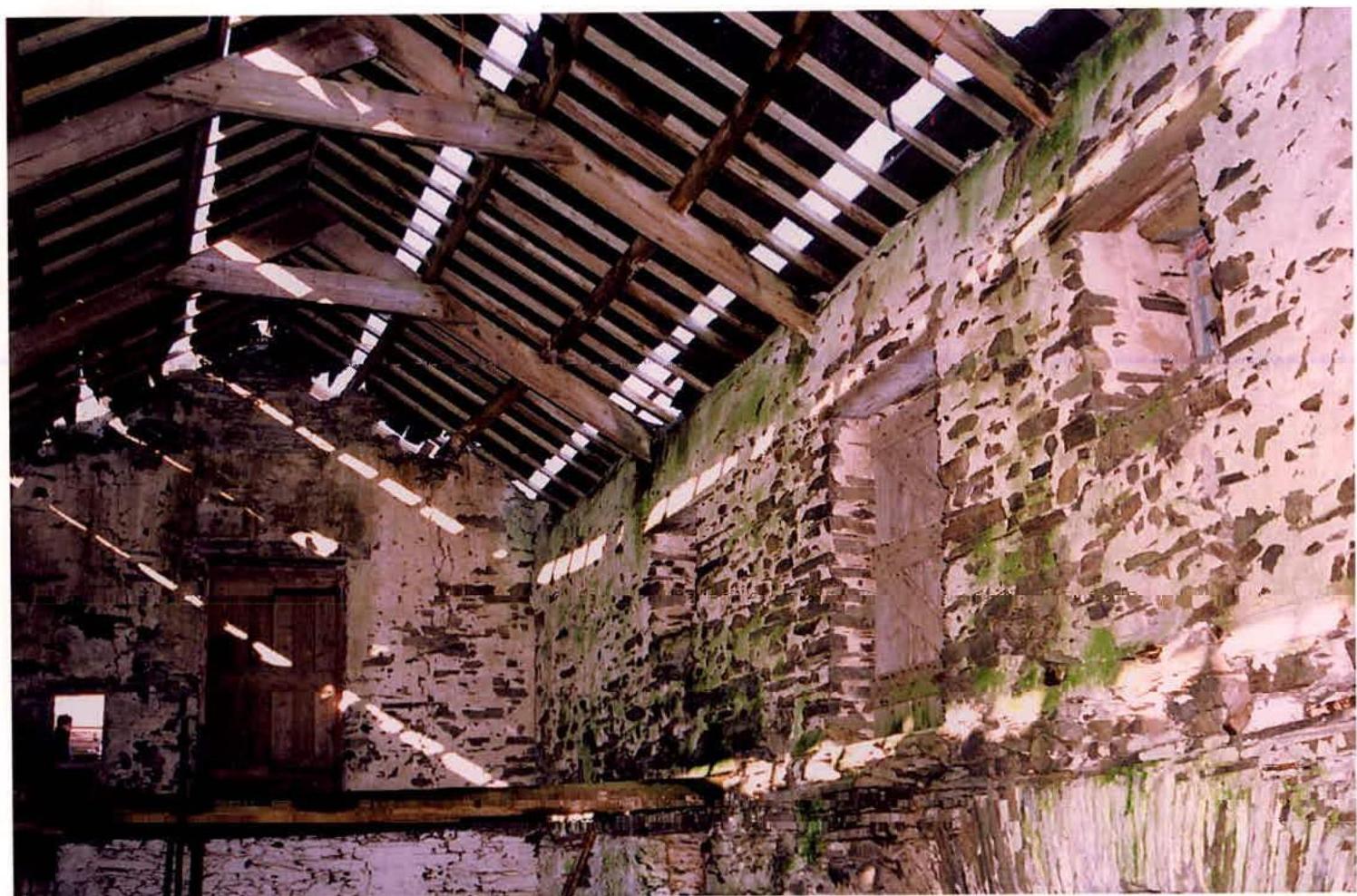 Interior photograph of a dilapidated stone building, likely a barn, showing exposed wooden roof beams and light entering through gaps in the roof structure.