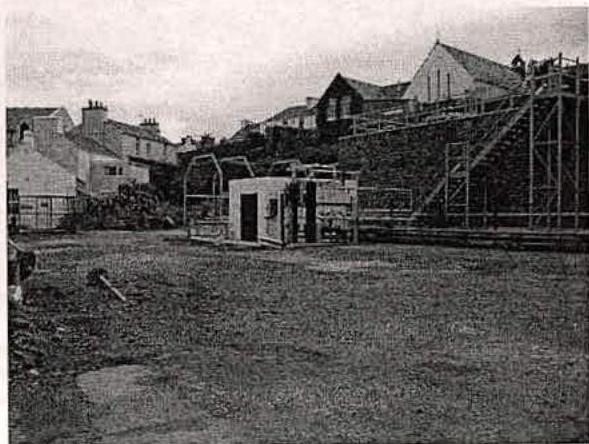 This black and white photograph shows a site area featuring a small white structure, fencing, and scaffolding on a building, with houses on a hill in the background.