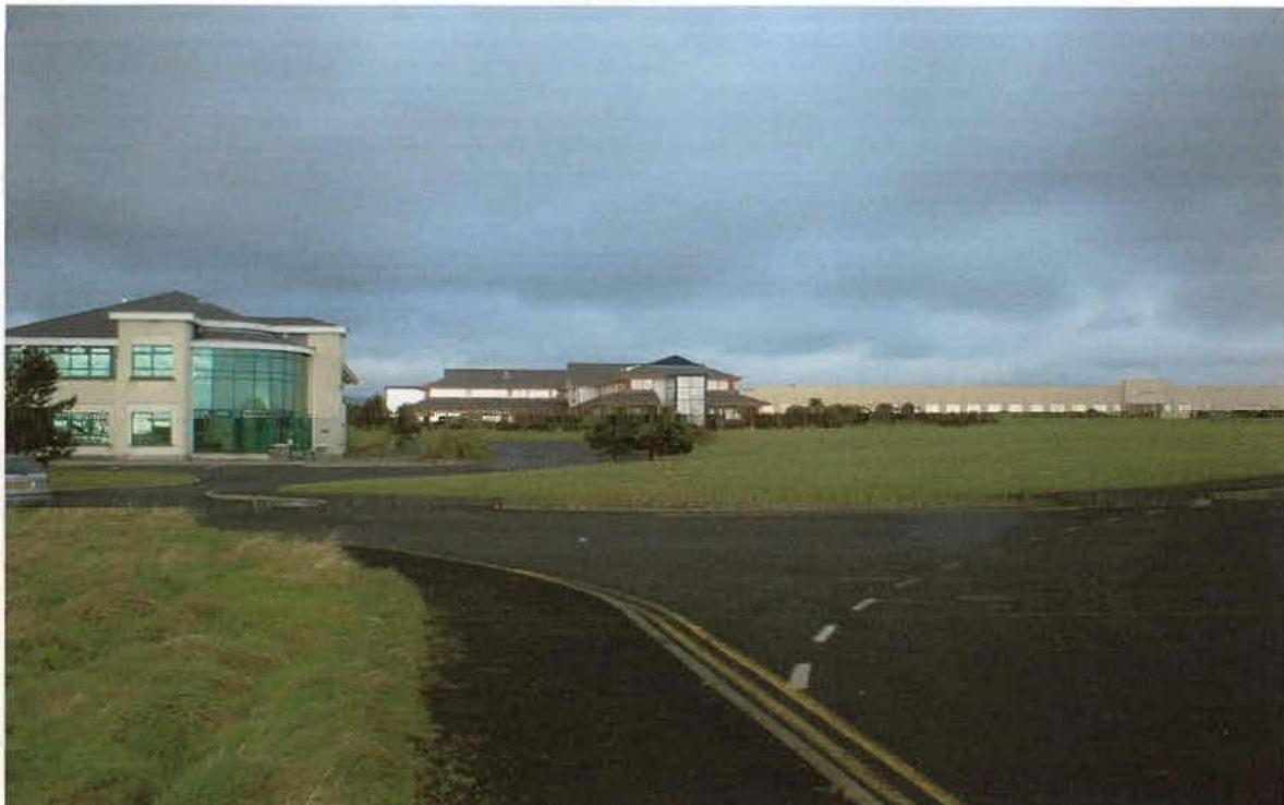 A photograph showing a modern office building with a curved glass facade on the left and a larger commercial complex in the background, viewed from a roadside.