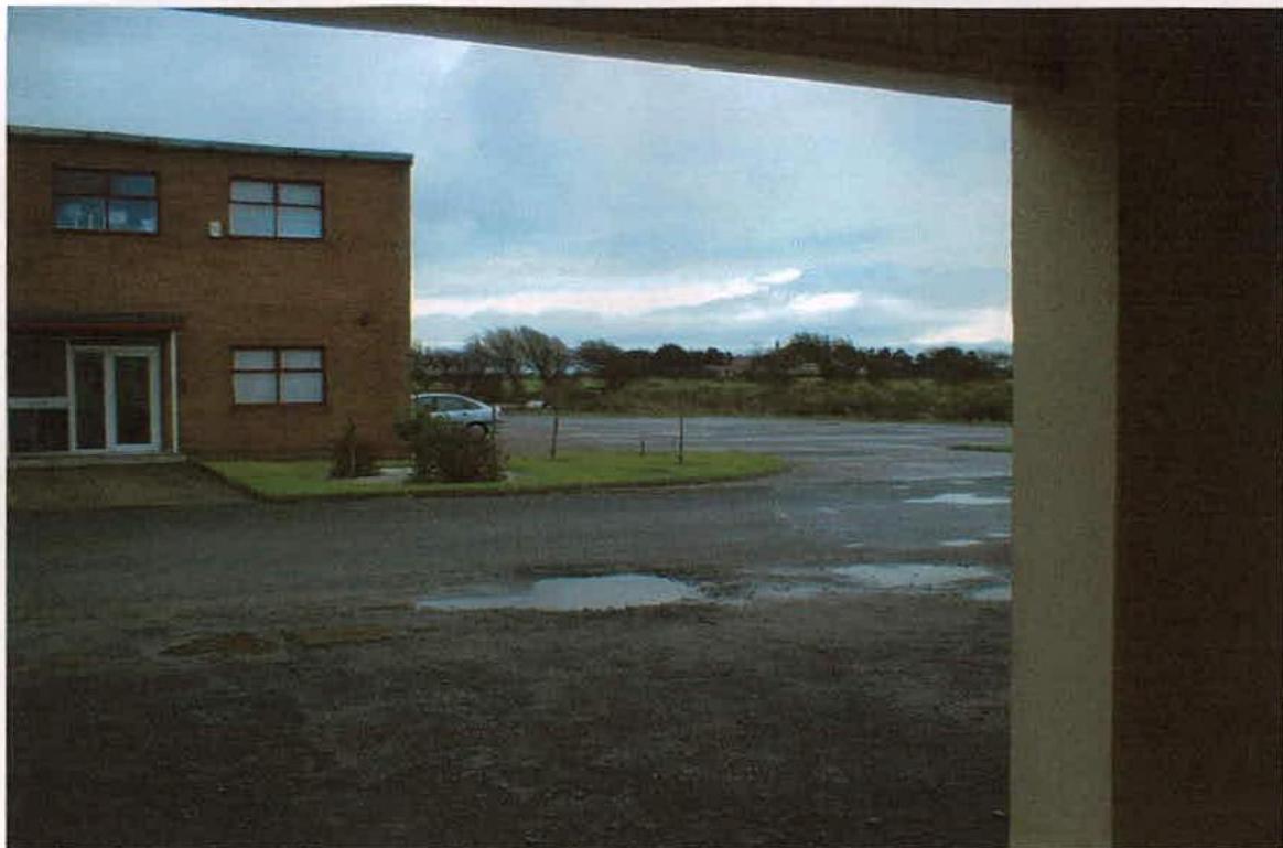 A photograph showing the exterior of a two-story brick office building with a paved parking area and open land in the background.
