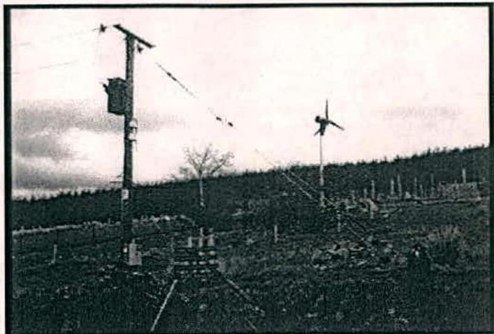 Grainy black and white photograph showing a rural hillside with a wind turbine in the distance and a utility pole in the foreground.
