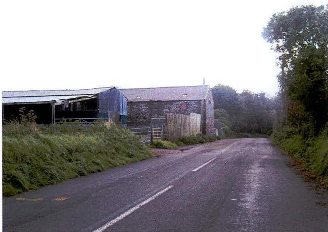A photograph showing a rural road with agricultural buildings on the left, including a stone barn and a metal shed.