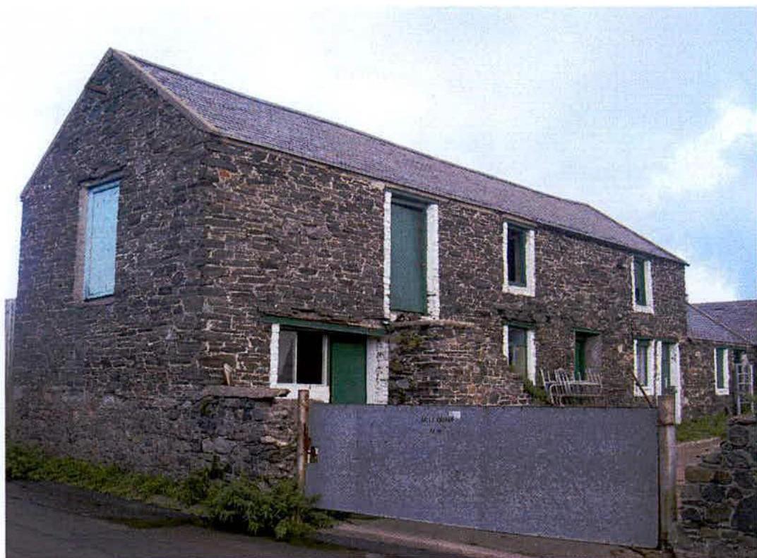 A photograph showing the exterior of a two-story stone farmhouse or barn with a slate roof and green shutters.