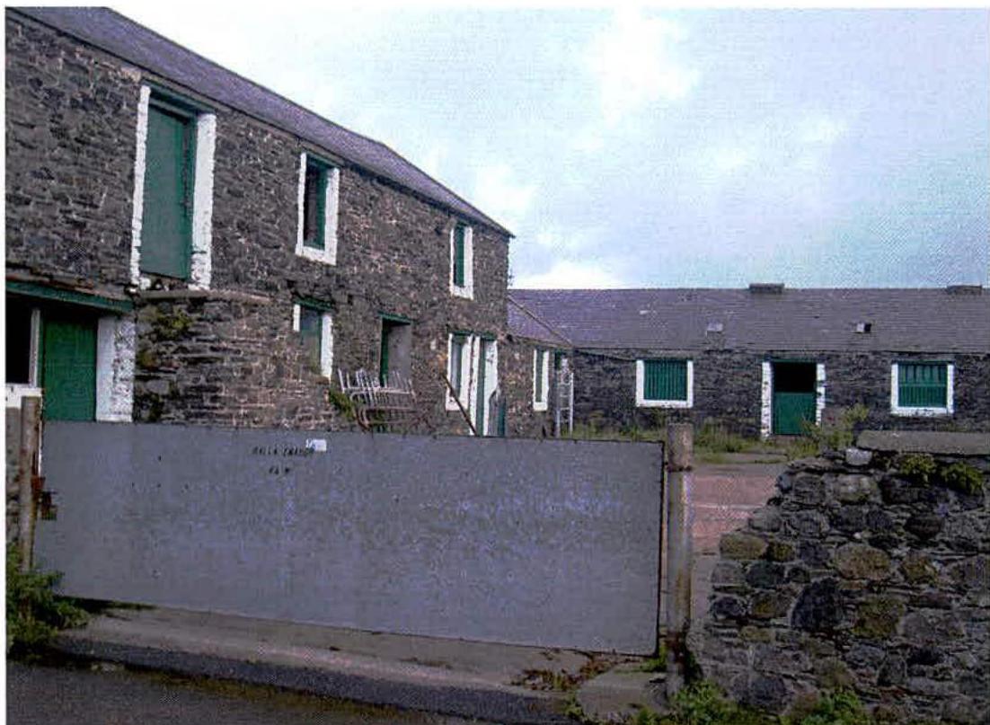A photograph showing a stone farmhouse and adjacent stone outbuilding with green doors, viewed through a large grey metal gate.