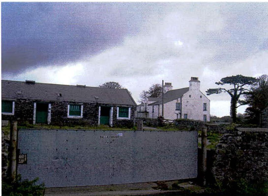 A photograph of a rural farmstead featuring a white two-story farmhouse and a long stone outbuilding situated behind a grey metal gate.
