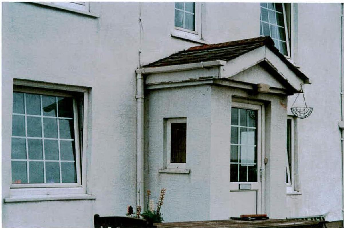 A photograph of the exterior of a white rendered residential building, featuring a small single-storey extension or porch structure with a door and window.