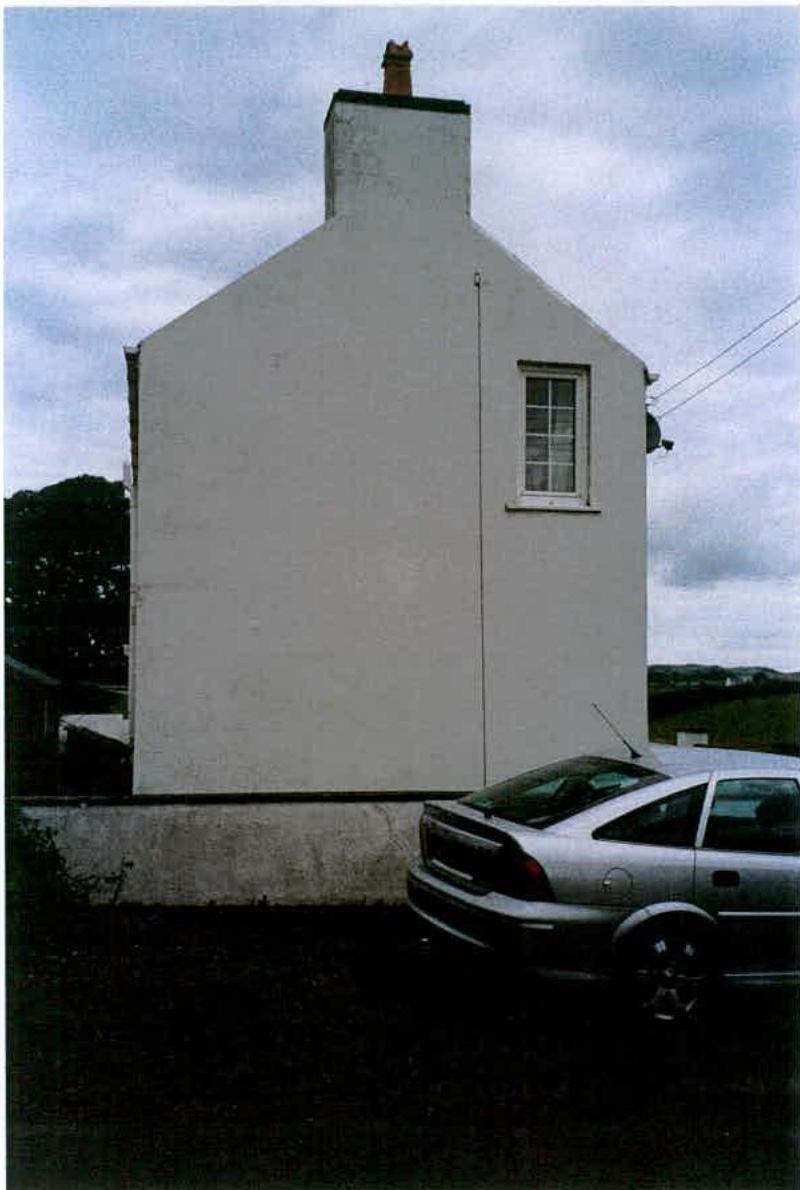 A photograph showing the rear elevation of a white, two-story detached house with a prominent chimney stack. A silver car is parked in the foreground on a dark surface, likely a driveway or hardstanding area.