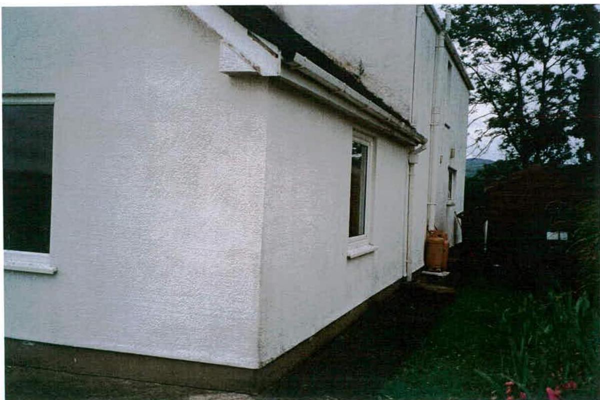A photograph showing the white rendered exterior corner of a detached house with windows and a garden area to the side.