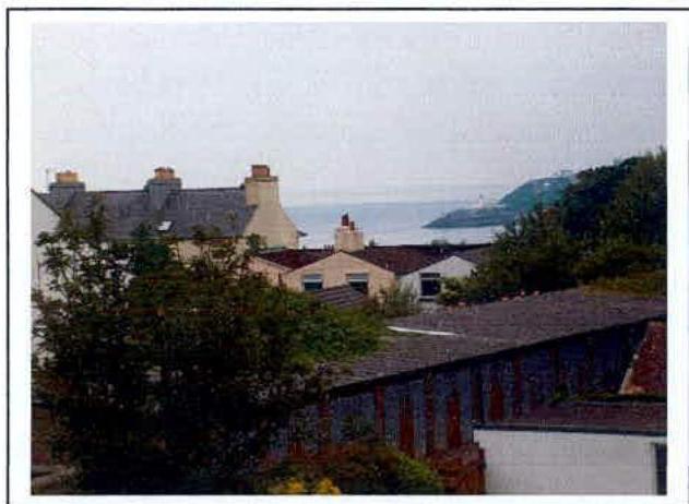 A photograph showing a view over residential rooftops and chimneys towards a coastal landscape with the sea in the background.