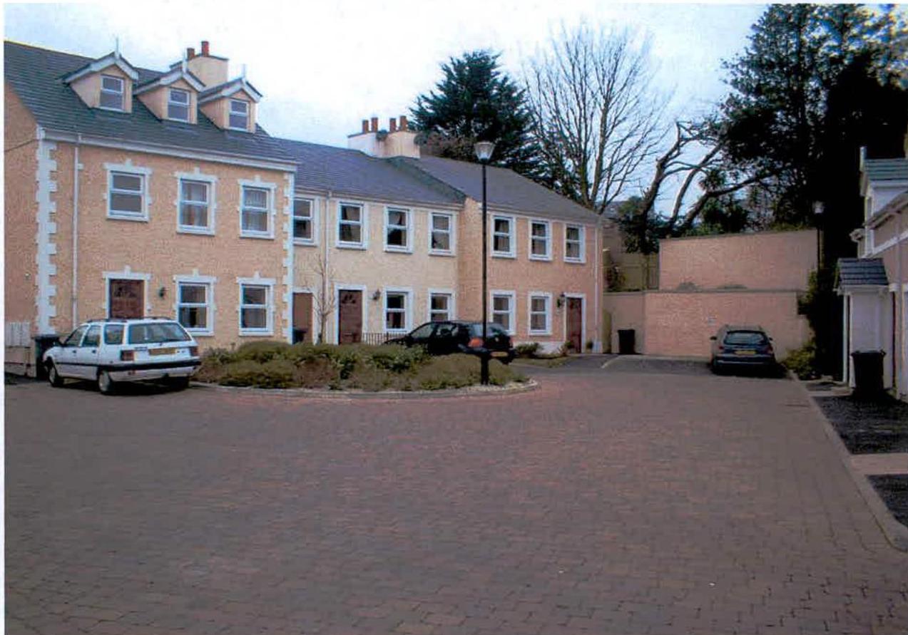 A photograph showing a row of two-story residential houses with cream facades and a large paved parking area in the foreground.