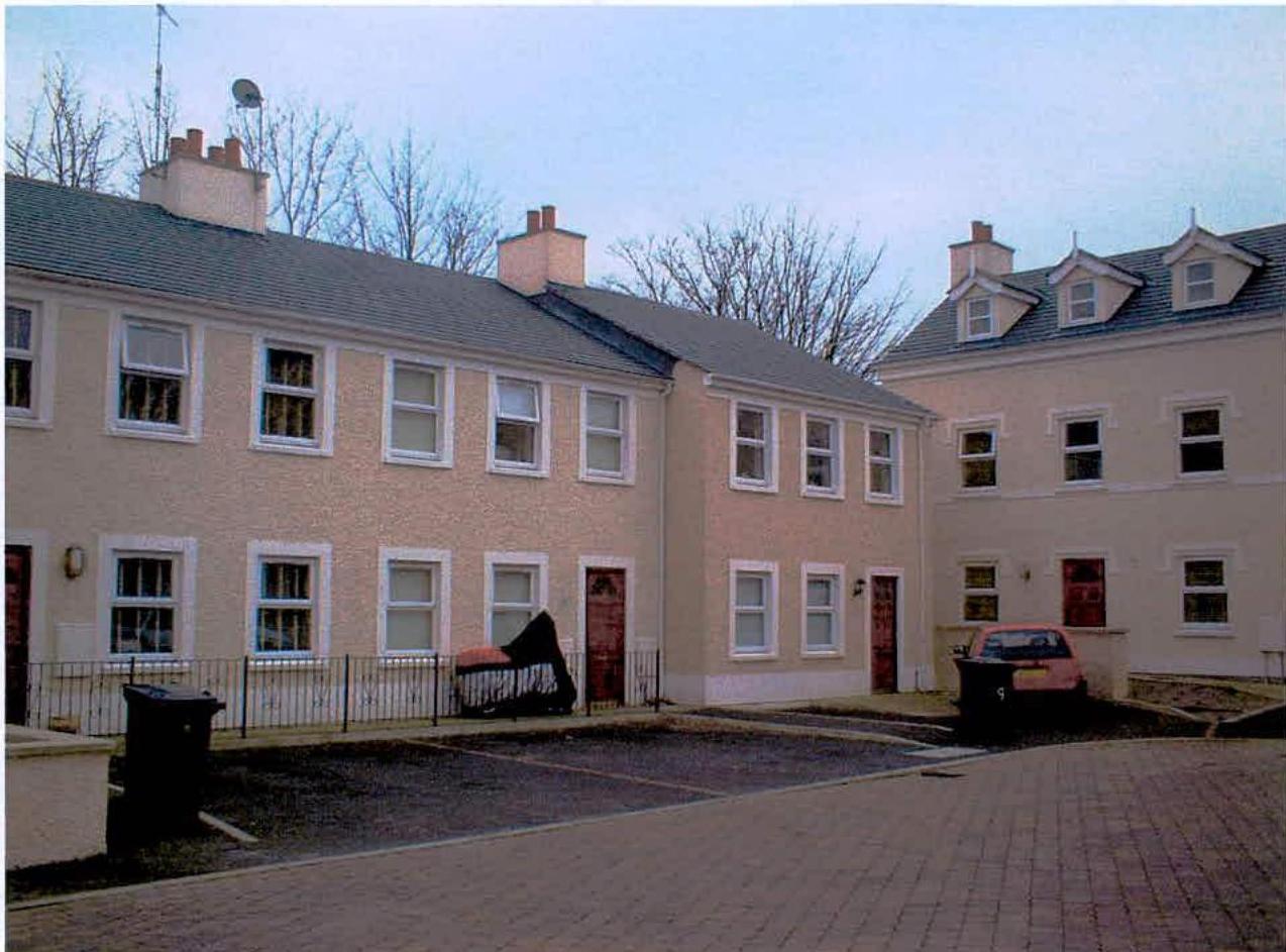 A photograph showing a row of cream-colored semi-detached houses with white window frames and slate roofs, featuring paved parking areas and parked cars in the foreground.