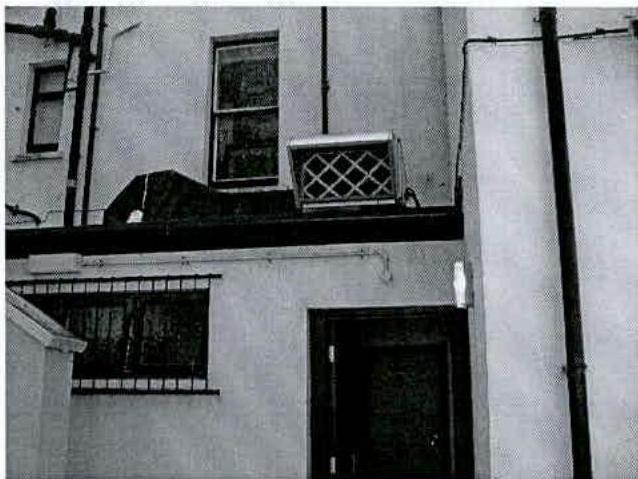 A black and white photograph showing the rear exterior of a building, featuring a ventilation unit mounted on the wall and vertical drainage pipes.