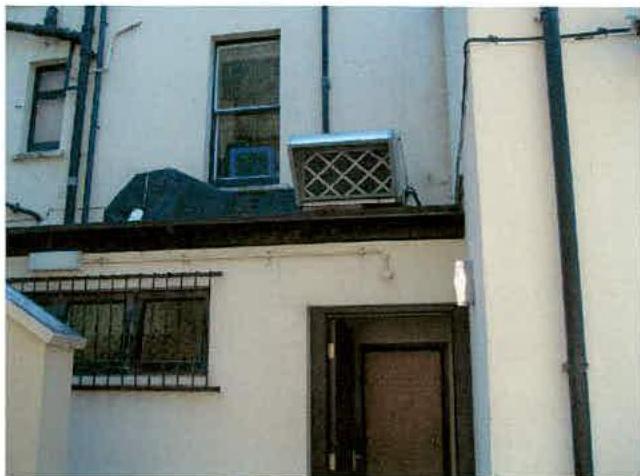 A photograph showing the rear exterior of a white building with a flat roof section featuring a metal ventilation unit. The image also displays downpipes, windows, and a door on the facade.
