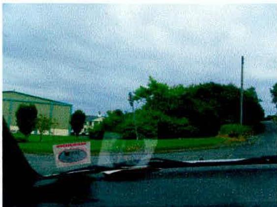 A grainy photograph taken from inside a vehicle showing a large industrial warehouse building and surrounding greenery. A sign is visible on the grass verge in the foreground.