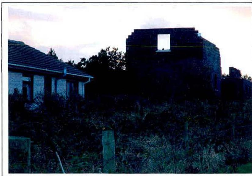 A dim photograph showing an existing single-story bungalow on the left and a dark, two-story block structure on the right, likely representing the proposed extension or new dwelling in an overgrown rural setting.