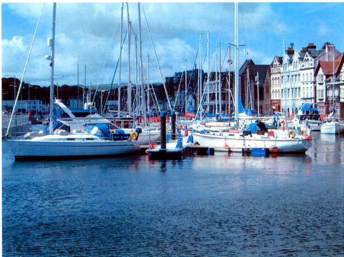 A photograph of a marina featuring several sailboats and yachts moored at floating pontoons. The background shows a row of multi-story buildings along the waterfront.