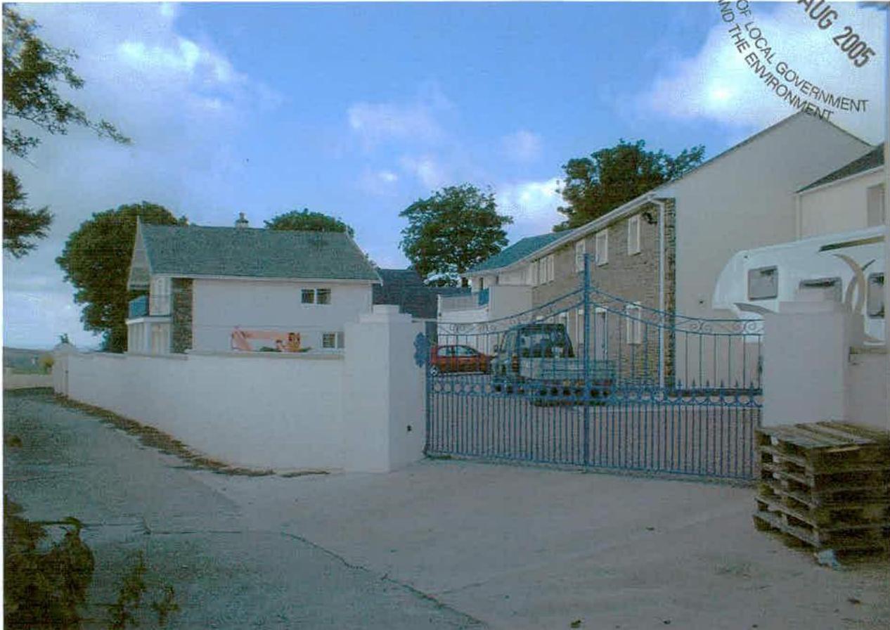A photograph showing a rural street scene with a white house, a blue metal gate, and a larger white building with a caravan parked nearby.