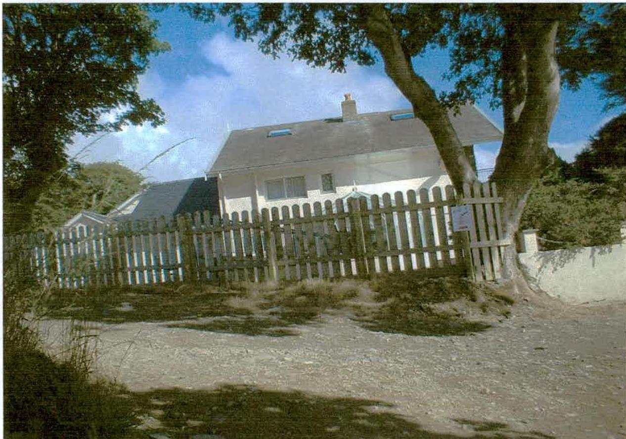 A photograph showing a white two-storey detached house with a pitched roof and skylights, situated behind a wooden picket fence in a rural setting.