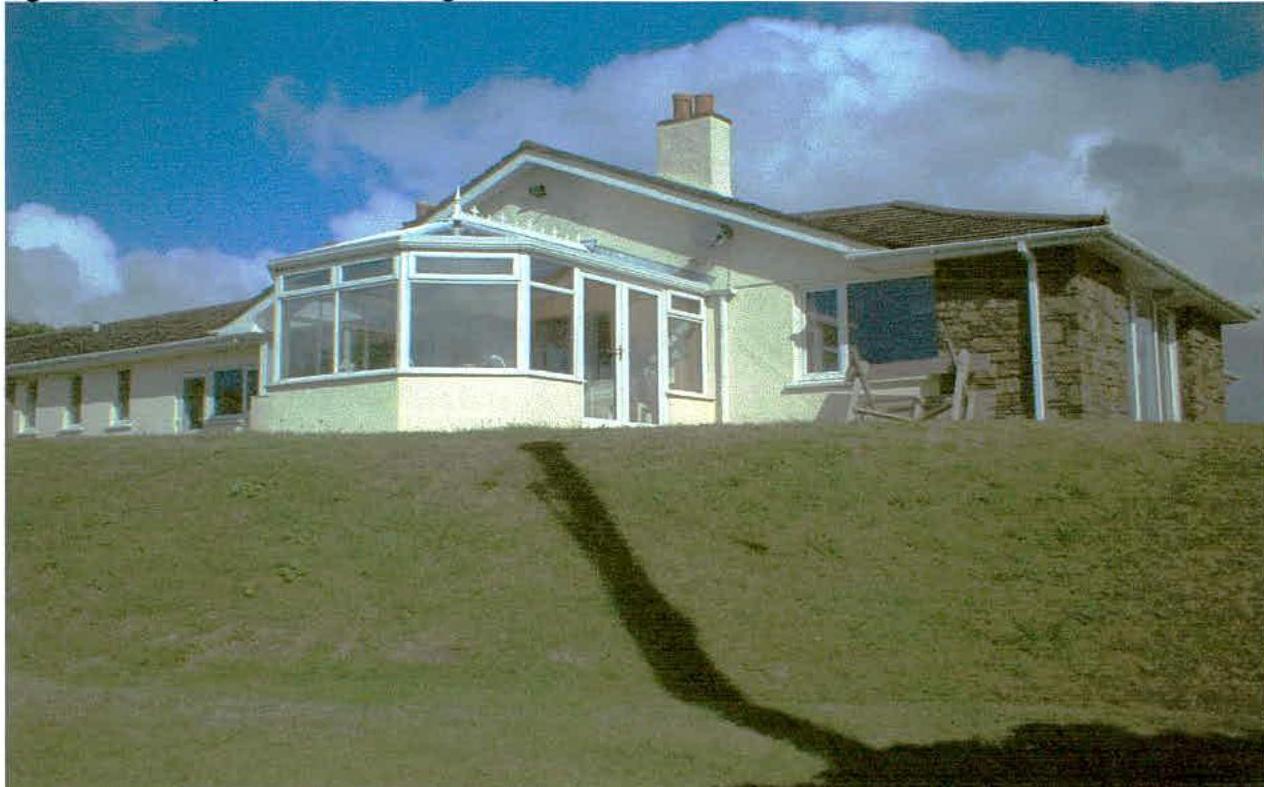 A photograph showing a single-story detached bungalow with a large conservatory extension situated on a grassy slope under a blue sky.