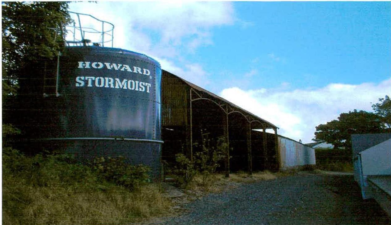 A photograph showing a large agricultural silo labeled 'HOWARD STORMOIST' alongside a long, open-sided metal barn structure on a rural site.