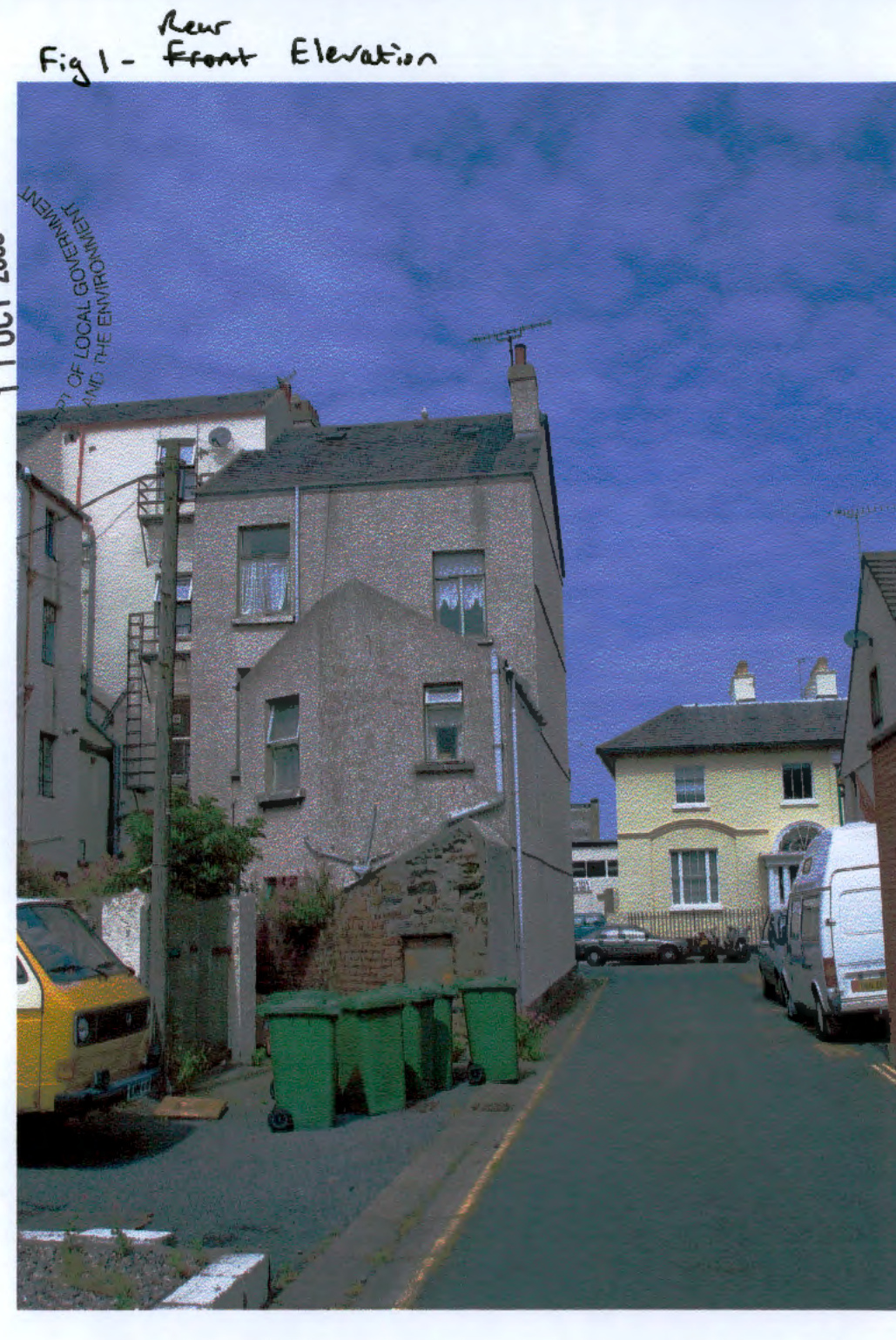 A photograph showing the exterior of a two-story residential building with pebbledash walls, situated on a street with parked vehicles and green waste bins. Handwritten text at the top reads 'Rear Fig 1 - Front Elevat...
