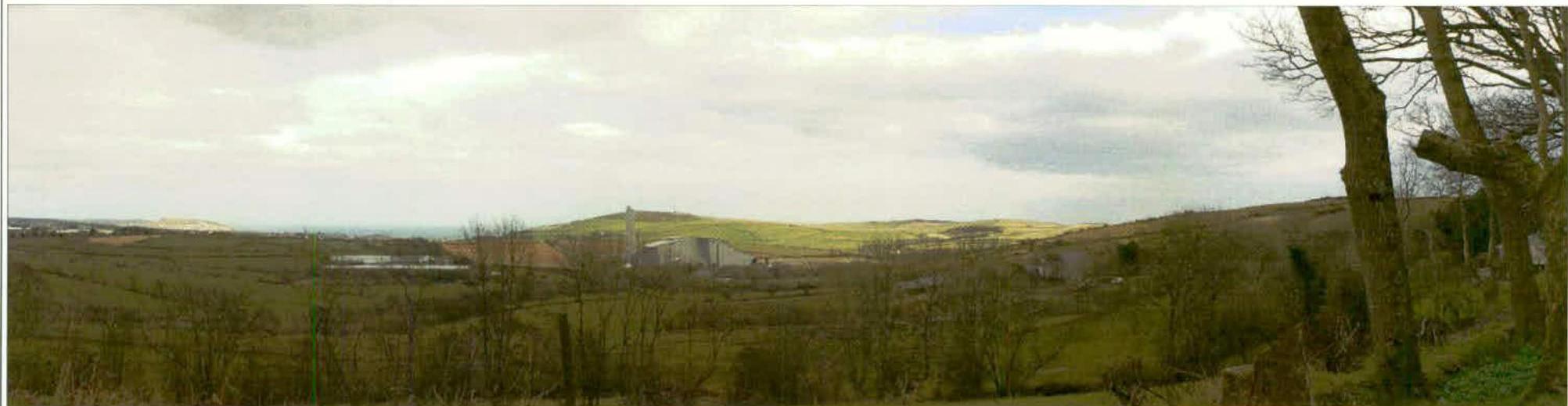 A panoramic photograph showing a rural landscape with rolling green hills and fields. Industrial-style buildings are visible in the middle distance, likely depicting the proposed facility site.