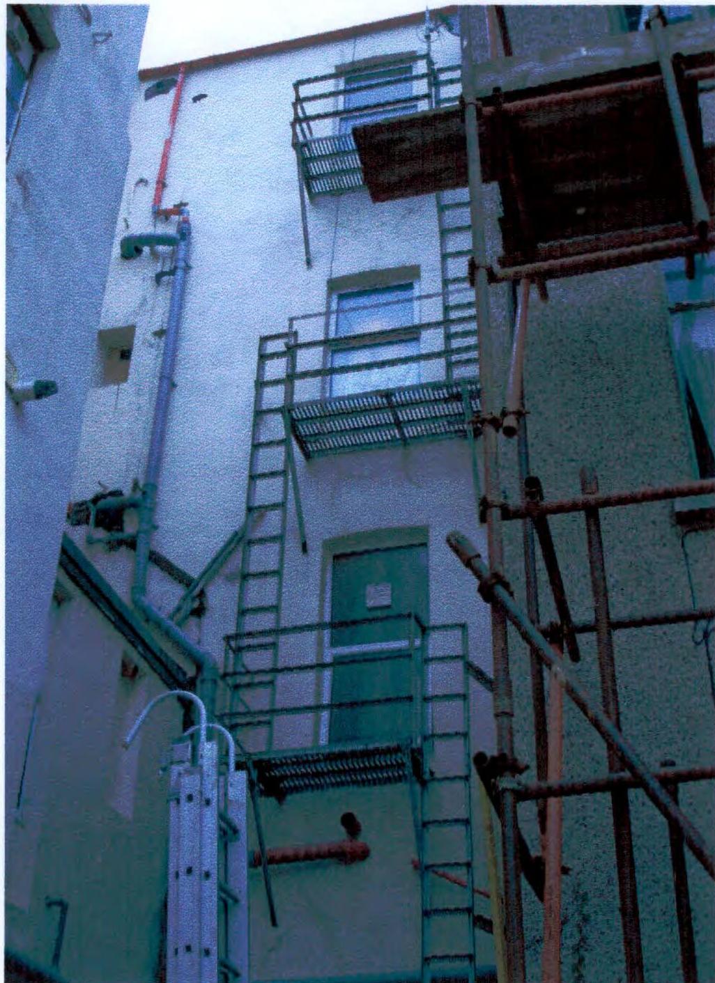 A grainy photograph showing the side or rear elevation of a white building featuring a green external metal fire escape staircase and vertical drainage pipes.