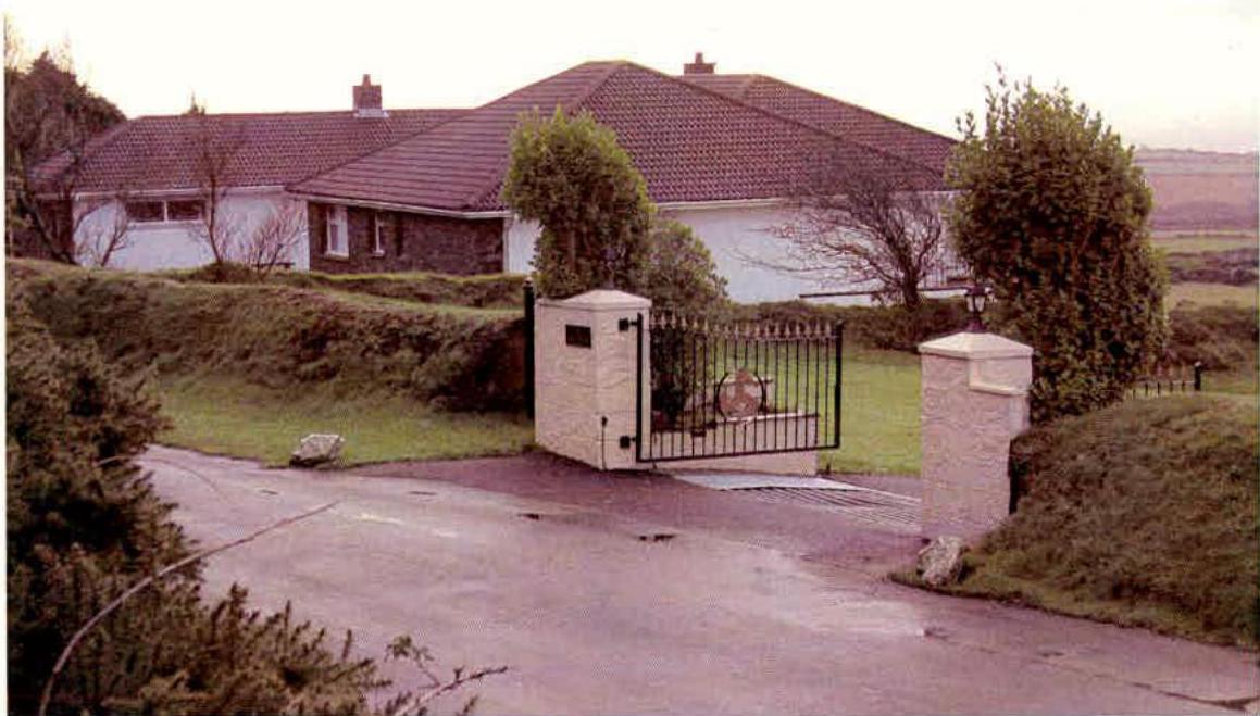 A photograph showing a detached single-story bungalow with a tiled roof, viewed from a driveway entrance featuring stone gate pillars.