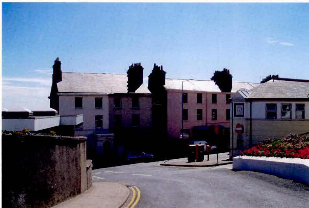 A street-level photograph showing a row of existing terraced residential buildings and a road junction, representing the site proposed for demolition and redevelopment.