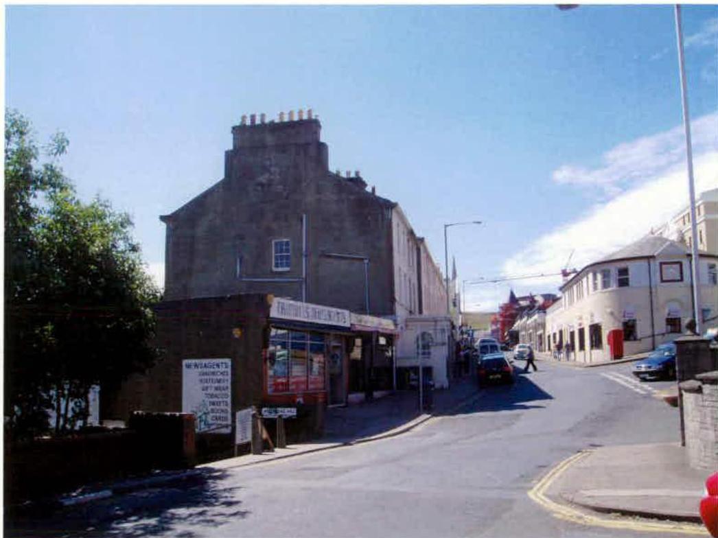 A street-level photograph showing an existing corner building with a shop front, likely the site proposed for demolition and redevelopment into offices and apartments.