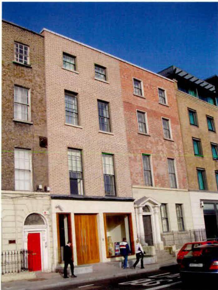 A street-level photograph showing a row of attached brick buildings, featuring a central light-colored structure with a modern ground floor flanked by older facades.
