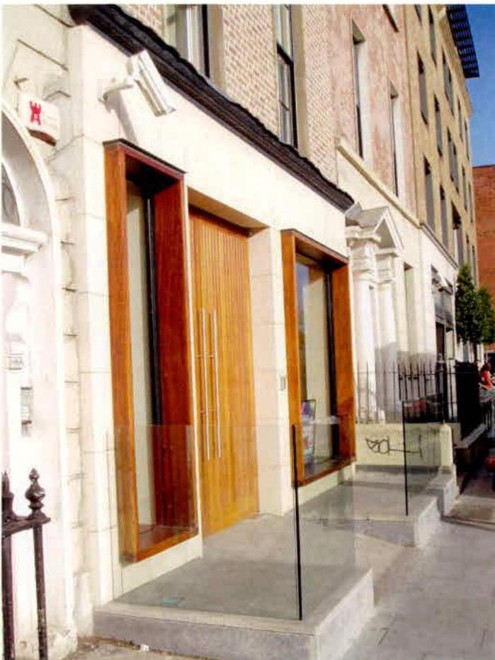 A street-level photograph showing the exterior facade of a building with large wooden doors and windows, featuring stone cladding and glass railings.