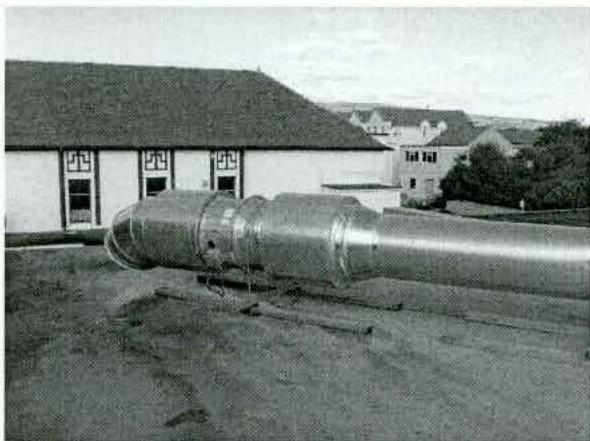 A black and white photograph showing a large metal flue pipe lying on the ground in the foreground with a building and hills in the background.