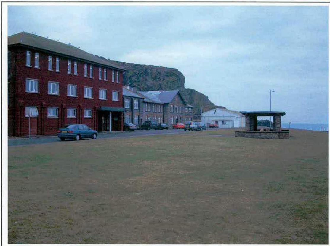 A photograph showing a street scene featuring a large red brick building and smaller stone cottages, set against a coastal cliff backdrop with a grassy foreground.